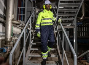 Trabajador industrial descendiendo escaleras metálicas con uniforme de alta visibilidad, casco, guantes y protección auditiva en planta de procesos.